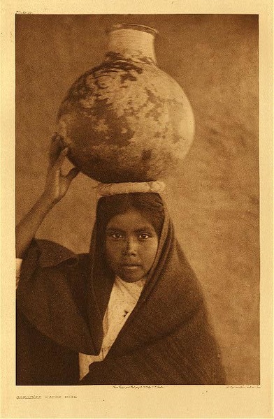 Edward S. Curtis - Plate 054 Qahatika Water Girl - Vintage Photogravure - Portfolio, 22 x 18 inches - Edward Curtis did not write a description for this photograph depicting a young Qahatika girl. She is carrying an earthen vessel on her head containing water and is dressed in a typical brown robe. The picture was taken in 1907. <br> <br>Curtis Grew up in Poverty and his formal education ended in 6th grade. There is a chance that he related to the Native Americans in a way and this is why he thought it so important to photograph them. Whatever his motives were we are fortunate to have these images of a race that may have been totally lost otherwise.