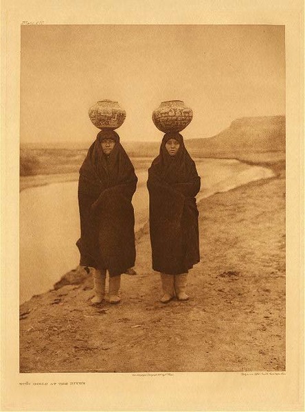 Edward S. Curtis - Plate 610 Zuni Girls at the River - Vintage Photogravure - Portfolio, 22 x 18 inches - In this picture by Edward S. Curtis, two Zuni girls carry water on their heads. They are adorned in long brown robes and in a desert like landscape. Seemingly young, one might guess the girls area round 15 years of age. The pots they carry on their heads are quite large and painted with symbols and imagery. Walking along the river, the two girls directly face the camera. They are surrounded by a soft, misty light, almost hiding the mesa in the background. <br> <br>This photogravure was taken by Edward Curtis in 1903 and printed on Japon Vellum Paper. The original vintage piece is now on display in our Aspen Art Gallery.