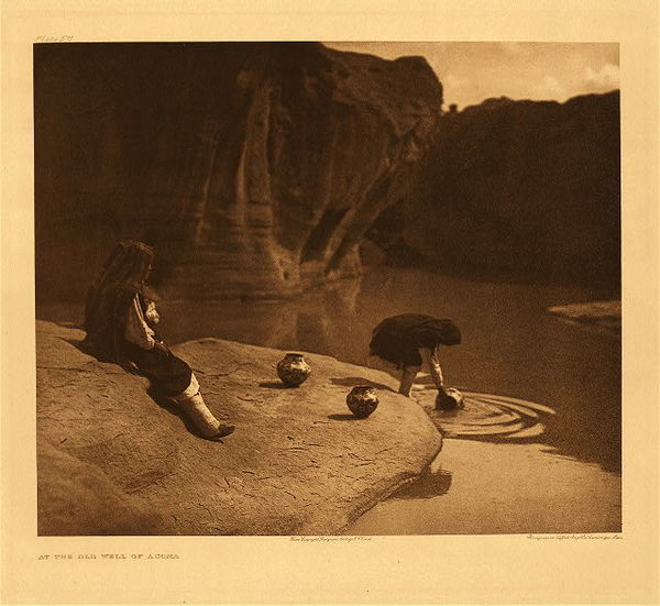 Edward S. Curtis - Plate 571 At the Old Well of Acoma - Vintage Photogravure - Portfolio: 18 x 22 inches - These young women are pictured collecting water on the rock of Acoma using beautiful earthen vessels. This specific spot was noted in 1540 by Coronado’s explorers as a place to collect snow and water. The soft ripples in the water, in contrast to the harsh and dominating rocks make a beautiful juxtaposition in this photogravure taken by Edward S Curtis.