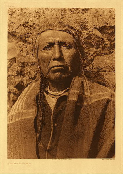Edward S. Curtis - Plate 282 Hlalakum - Wishham - Vintage Photogravure - Portfolio, 22 x 18 inches - Here is a strong, close-up portrait of Wisham individual, brow furrowed and mouth slightly downturned. The individual's proud posture is softened by a blanket cloaking the body and a braid falling alongside their shoulder. The emotion of this piece is palpable, accentuated by the sitter's direct gaze. <br> <br>"The myths of the Wishham are exceedingly interesting in that they show unusual wealth of imagination and vivacity, yet they are disappointing in their incomplete cosmology and their inattention to obvious phenomena. The Wishham attainment of supernatural attributes was attended with far less than ordinary travail. When seven or eight years of age a boy was instructed by his father or other male relative in the mysteries of the acquirement of "yuhlmah," and bidden to begin his journey to lonely spots among the high hills, that the spirits might come to him." from Edward S. Curtis' "The North American Indian", Volume VIII