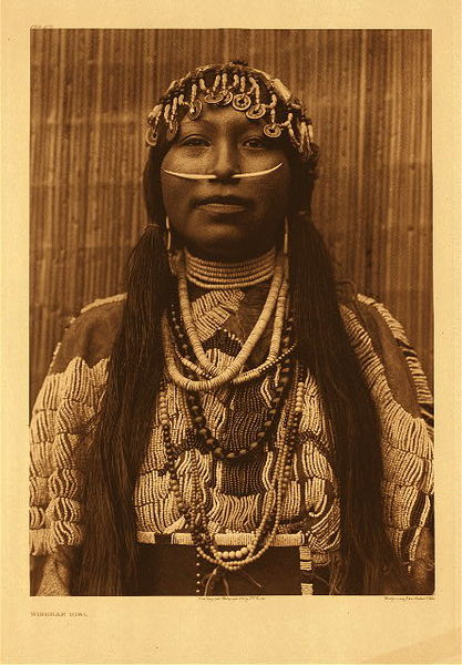 Edward S. Curtis - Plate 278 Wishham Girl - Vintage Photogravure - Portfolio, 22 x 18 inches - The subject is clothed in a heavily beaded deerskin dress of the plains type. The throat is encircled by strands of shell beads of native manufacture, heirlooms which were obtained by the original Wishham possessor from the Pacific slope. Pendant on the breast are strands of larger beads of the same kind, as well as of various kinds brought into the country by the traders of the Hudson's Bay Company. An indispensable ornament of the well-born person was the dentalium-shell thrust through a perforation in the nasal septum; occasionally, as in this case, two such shells were connected by means of a bit of wood pushed into the hollow bases. Tied to the hair at each side of the face (see the following plate) is another dentalium-shell ornament, which is in reality an ear pendant transferred from the lobe of the ear (where its weight would be inconvenient) to the hair. The head-dress consists of shells, shell beads, commercial beads, and Chinese coins. The coins made their appearance in the Columbia River region at a comparatively early date. This form of head-dress was worn on special occasions by girls between the age of puberty and their marriage.” – Edward Curtis