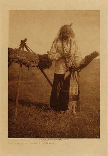 Edward S. Curtis - Elk Head and Sacred Pipe Bundle - Vintage Photogravure - Volume, 12.5 x 9.5 inches - This photo was taken by Edward S. Curtis in 1907 and the story is as follows: According to the Lakota Legend they were a people with little knowledge of how to live or worship before the coming of a divine messenger named “White Buffalo Woman”. White Buffalo Woman left with her people along with her teachings a sacred calf pipe. At the time of Edward S. Curtis’ photograph Elk Head had been the keeper and protector of the pipe for 31 years with 6 other keepers prior to him. This myth is of relatively recent origin, about 368 years before Curtis met the tribe. This amount of time would have meant that Elk Head’s predecessors had protected the sacred pipe for about 56 years each. To the Lakota the pipe is the holiest of things. <br> <br>Elk head holds the beaded and quilled pipe bag in this image while standing in the plains. The image was taken for Curtis’ volume three and was printed on Dutch Van Gelder. It is now available for sale in our Aspen Art Gallery.