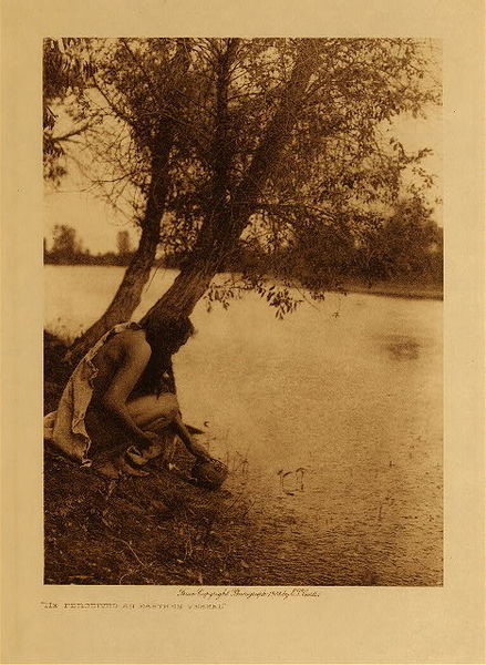 Edward S. Curtis - He Perceived an Earthen Vessel - Vintage Photogravure - Volume, 12.5 x 9.5 inches - This Edward Curtis photo was taken in 1908 and depicts a reenactment of the Legend of Taking up the Bowl. <br> <br>The Hidatsa tribe had a great ceremony in addition to the Sun Dance called the Ceremony of the Bowl. According to the legend a long time ago one of the tribe was fasting on a shore far from the village and crying to the spirits to pity him. Just before the sun went down he saw something on the shore where the waves had lapped the sand and he found it to be an earthen vessel marked with the track of a brant around its rim. <br> <br>He took it home and that night he had a vision. The bowl spoke to him saying, "My child, I am Old Woman Who Never Dies. Hold me sacred, and I will bring you good fortune, for I have many friends among the spirits. The corn and buffalo-paunch are my food. I shall teach you the songs and rites of a ceremony that will cause your people to prosper and bring rain upon your crops. Make offerings to me of buffalo-paunches, and hang them before me on cottonwood stakes. Prepare a pipe and tobacco for Itsihkawahidish and Adhapushish, who aid me in making medicine, for they are men and like to smoke. All the birds and animals living on this lake are of my medicine. Let no man who has blood on his hands enter the lodge where this ceremony takes place, nor permit any woman to be present." <br> <br>She revealed to him the rites of taking up the bowl. This would be a huge ceremony for the tribe. <br> <br>This image was printed on Dutch Van Gelder paper and is available for sale in our Aspen Art Gallery.
