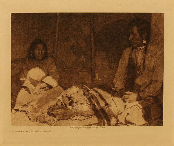 Edward S. Curtis - *50% OFF OPPORTUNITY* A Beaver Bundle - Blackfoot 50% off opportunity - Vintage Photogravure - Volume, 9.5 x 12.5 inches - This image represents the very important ceremony surrounding the planting of tobacco. Two men sit beside each other holding the beaver bundles that are likely full of tobacco seed. They are in the shadows and the light shines mostly on the bundles. This photogravure was taken by Edward S. Curtis and is now on display in our Aspen Art Gallery. <br> <br>“The principal Beaver Bundle was shown the writer in a tent at a haying camp. It lay in the place of honor on a pile of folded rawhides. It was wrapped in a tanned buffalo-skin colored red, and was securely wound with plaited rawhide rope. Under the turns of the rope were two hardwood dibbles, each having the incised outline of a beaver on its flat side. The handle ends were ornamented with beads. With the sticks were two long pipes separately wrapped in red flannel. <br> <br>The ceremony of planting tobacco is held in great awe. The following account, apparently half myth and half legend, purports to explain the origin of the beaver bundles and ritualistic tobacco planting.” -From Page 184 in volume 18 of Edward Curtis' North American Indian