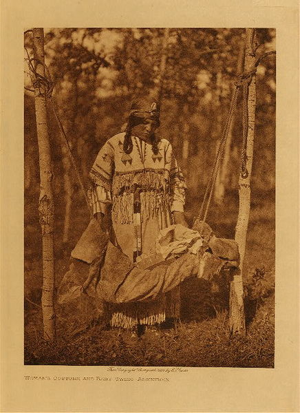 Edward S. Curtis - Woman's Costume and Baby Swing - Assiniboin - Vintage Photogravure - Volume, 12.5 x 9.5 inches - This image by Edward Curtis depicts a, Assiniboin mother in an elaborately decorated dress swinging her young baby in a hammock-like swing. The swing is tied between two trees. A serene image of the beauty of motherhood in the Native tribes. <br> <br>This image was taken in 1926 by photographer Edward S. Curtis and was printed on Japon Vellum. The Original photogravure is available for sale in our Aspen Art Gallery.