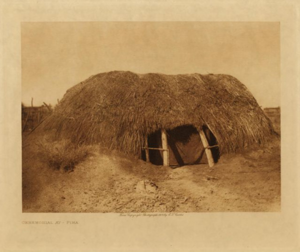 Edward S. Curtis - *50% OFF OPPORTUNITY* Ceremonial Ki - Pima - Vintage Photogravure - Volume, 9.5 x 12.5 inches - The early dwellings of the Pima and their immediate congeners were quite alike: a dome-shape structure about seven feet high and fifteen feet in diameter at the base. A circular excavation twelve to eighteen inches deep was first made, in the centre of which four crotch posts were set about five feet apart, forming a square. Two heavy roof timbers rested upon these posts, five feet from the ground, supporting ten or more stout cross-beams. Numerous stave-like ribs of mesquite, tied to horizontal poles that extended around the outside like hoops, were stretched from the roof timbers to the bottom of the shallow excavation. The whole was thatched with arrow-brush and covered with clay, leaving only a small opening at the base on the eastern side as an entrance. Only a few of the old form of houses are now in use among the Pima. For the greater part their domiciles are rectangular, with flat roofs and straight walls of mud filled in between poles fastened horizontally to opposite sides of stout posts, or with brush-wattled walls plastered inside and out with mud.