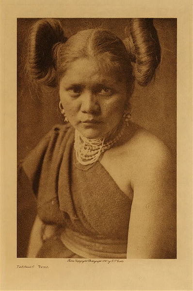 Edward S. Curtis - Tsetsanu (Tewa) - Vintage Photogravure - Volume, 12.5 x 9.5 inches - The young woman “Tsetsanu” from the Tewa is pictured here by Edward S. Curtis. Tewa was a subset of the Hopi and had similar characteristics and dress. Her hair is done in whorls, indicating that she in unmarried. The subject is dressed stylishly with jewelry and an off the shoulder dress. <br> <br>This image was taken in 1921 by photographer Edward S. Curtis and was printed on Japon Vellum. The Original photogravure is available for sale in our Aspen Art Gallery.