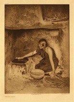 Edward S. Curtis - Plate 432 The Piki Maker - Vintage Photogravure - Portfolio, 22 x 18 inches - This young Hopi maiden is pictured here making piki, a cornbread that is cooked by spreading the batter upon the baking stone with the bare hand. This small baking area just fits one person and she has a basket on her left side for collecting the baked pieces of bread. The subject’s hair has been twisted on either side of her head and she is wearing a simple long brown cloth dress.
<br>
<br>“Piki is cornbread baked in colored sheets of paper-like thinness. The batter is spread on the baking stone with the bare hand, and the quickly baked sheet is folded and laid on the basket at the baker's left.” - Edward Curtis