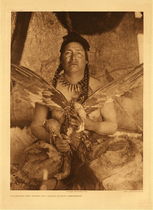 Edward S. Curtis - *50% OFF OPPORTUNITY* Plate 634 Placating the Spirit of a Slain Eagle - Assiniboin - Vintage Photogravure - Portfolio: 22 x 18 inches - This middle aged Assiniboin Man holds the spread winged carcass of an eagle. Eagles were coveted for their feathers which were used in many ways. Some of these uses include ornaments and fetishes. Once caught, the sacred eagle was honored by an elaborate ceremony that took place to placate the eagle’s spirit. In this image the man is participating in this ceremony. 
<br>
<br>The subject is likely the one who has slain the eagle. He wears a necklace made of claws and two long braids. He also carries some accoutrement to assist in the ceremony. This picture was taken by Edward Curtis in 1926 and is on display in our Aspen Art Gallery.
<br>
<br>“Maintaining respectful, reciprocal relationships with slain animals was vital to the everyday lives of native peoples because the ability to connect with the spirit world was important. 
<br>“The path to power provided several options. Working alone, individuals might perform a vision quest, make tobacco offerings, or sing special songs to spirits of the animal and fish. After killing an animal, the hunter respectfully thanked its spirit for the sacrifice and tried to treat its body with respect.”
<br>
<br>Edward Curtis recognized the immense value in the word “respect” within the pages of The North American Indian, and the photogravure portraits. Any cultural information in the pages of his volumes was obtained with permission of the tribes he wrote about.