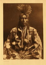 Edward S. Curtis - Plate 208 A Piegan Dandy - Vintage Photogravure - Portfolio, 22 x 18 inches - One of the most striking subjects in the entire Edward Curtis collection. 
<br>The Sun Dance or “Okun” - This costume may be prominent in the ceremony for the Piegan the principal deity; the sun or "Napiw," who was called upon for supernatural aid. 
<br>
<br>As with many North American Indian tribes, the sun was the main deity. Many celestial bodies are here to focus of “supplication in Prayer by any individual who desired supernatural aid, whether or not he or she had ever seen in a vision the one so addressed.”
<br>
<br>The influence of tribes upon each other was witnessed by Edward Curtis as he learned of similar rituals.