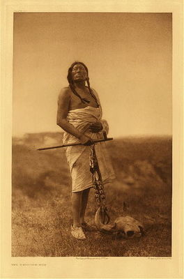 Edward S. Curtis - Plate 076  The Medicine-Man - Vintage Photogravure - Portfolio, 22 x 18 inches - “Invocation and supplication enter so much into the life of the Indian that this picture of the grim old warrior invoking the Mysteries is most characteristic. The subject of the illustration is Slow Bull, whose biography is given in Volume III, page 189.” – Edward Curtis
<br>
<br>“All things passing understand are “waka.” When supplicating Waka-ta ka, the Indian conceives the Mystery as possessing and being all things that transcend his comprehension. After invoking successively each deity in his belief, he comprehends all in prayer, “Great Mystery!” and in the cry he has included all the forces of the universe, from that represented by the personal fetish on his body to the undefined consciousness of the infinite.” Volume III p 60
<br>
<br>Slow Bull was born in 1844 and participated in his first war-party at fourteen, engaging in fifty-five battles over his lifetime. At seventeen he captured one hundred and seventy horses from the Apsaroke and “In the same year he received medicine from buffalo in a dream while he slept on a hilltop, not fasting, but resting from travel on the war-path.” 
<br>
<br>He was 63 in this photogravure and had been the subchief of the Ogallala tribe for 30 years.