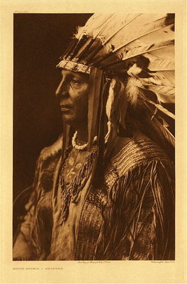 Edward S. Curtis - Plate 152 White Shield - Arikara - Vintage Photogravure - Portfolio, 22 x 18 inches - “A mixed-blood member of the medicine fraternity.”
<br>
<br>Spend a moment to look at the details in this extraordinary tunic.
<br>
<br>This Arikara wears an elaborate shirt, with meticulous details in this and the headdress. The details in the weaving are significantly different than designs in other Indian clothing.
<br>
<br>What we find in Arikara clothing is most like what we remember in many pictorial descriptions of the North American Indian; breechcloths, fringed buckskin tunics shirts and leggings. For protection from rain and cold a warm, buffalo robe was worn. When dressed for ceremony they wore elaborately decorated shirts and war bonnets with eagle feathers and beadwork, which symbolize courage and accomplishment. 
<br>
<br>"The Arikara are a semi-agricultural people, and have apparently been such from an early period, many myths and ceremonies bearing relation to growing corn and other products of the soil. The variety of corn raised is characteristic of the agricultural tribes in the Southwest-dwarf-like but very leafy stalks with many small ears of varicolored grains growing near the ground."
<br>
<br>"The basket common to these three tribes is claimed by the Arikara as originally a product of their own art, and writers acquainted with the basketry of southern Caddoan tribes assert this is a Caddoan pattern."
<br>            
<br>"The soul, called "Sishu," is responsible for all acts of man during life: it resides in the breast, and appears in the spoken word, in the look of the eye, in the movement of the muscles. It is the Sishu that rattles in the throat of the dying in an attempt to escape… All animals have Sishu, but not trees and inanimate objects."
<br> 
<br>"The Arikara developed the legerdemain of their all summer medicine ceremony to such an extent that other tribes, far and near, learned of their wonderful and potent magic. The superstition and credulity of the Indian are such that medicine-men living afar, as well as the tribesmen of the performers, believed these tricks to be the mysterious tricks of supernatural powers.