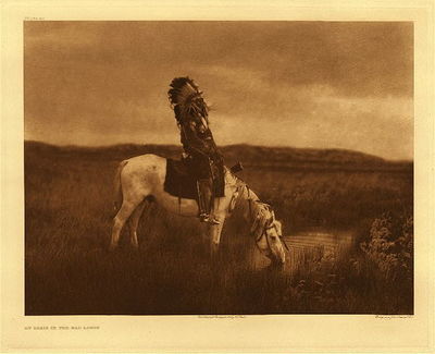 Edward S. Curtis - Plate 080 An Oasis in the Badlands - Vintage Photogravure - Portfolio, 18 x 22 inches - Born 1854. First war-party in 1865 under Crazy Horse, against troops. Led an unsuccessful war-party at twenty-two against Shoshoni. First coup when twelve horse-raiding Blackfeet were discovered in a creek bottom and annihilated. Led another party against Shoshoni, but failed to find them; encountered and surrounded a white-horse troop. From a hill overlooking the fight Red Hawk saw soldiers dismount and charge. The Lakota fled, leaving him alone. A soldier came close and fired, but missed. Red Hawk did likewise, but while the soldier was reloading his carbine he fired again with his Winchester and heard a thump and "O-h-h-h!" A Cheyenne boy on horseback rushed in and struck the soldier, counting coup. Engaged in twenty battles, many with troops, among them the Custer fight of 1876; others with Pawnee, Apsaroke, Shoshoni, Cheyenne, and even with Sioux scouts.
<br>Red Hawk fasted twice. The second time, after two days and a night, he saw a vision. As he slept, something from the west came galloping and panting. It circled about him, then went away. A voice said, "Look! I told you there would be many horses!" He looked, and saw a man holding green grass in his hand. Again the voice said, "There will be many horses about this season;" then he saw the speaker was a rose-hip, half red, half green. Then the creature went away and became a yellow-headed blackbird. It alighted on one of the offering poles, which bent as if under great weight. The bird became a man again, and said, "Look at this!" Red Hawk saw a village, into which the man threw two long-haired human heads. Said the voice,"I came to tell you something, and I have now told you. You have done right." Then the creature, becoming a bird, rose and disappeared in the south. Red Hawk slept, and heard a voice saying, "look at your village!" He saw four woman going around the village with their hair on the top of their heads, and their legs aflame. Following them was a naked man, mourning and singing the death-song.  Again he slept, and felt a hand on his head, shaking him, and as he awoke a voice said, "Arise, behold the face of your grandfather!" He looked to the eastward and saw the sun peeping above a ridge. The voice continued, "Listen! He is coming, anxious to eat." So he took his pipe and held the stem toward the rising sun. This time he knew he was not asleep, or dreaming: He knew he was on a hill three miles from the village. A few days later came news that of five who had gone against the enemy, four had been killed; one returned alive, and followed the four mourning wives around the camp singing the death-song. Still later they killed a Cheyenne and an Apsaroke scout, and the two heads were brought into camp.