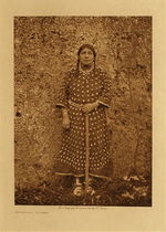 Edward S. Curtis - *50% OFF OPPORTUNITY* Apsaroke Woman - Vintage Photogravure - Volume, 12.5 x 9.5 inches - This Apsaroke woman stands facing the camera so that we can view her entire figure. She is wearing a dress very typical of the Apsaroke people generally made of sheep skin. On the neck and sleeves this dress may be embroidered with porcupine quills or beads but the most striking element of these dresses are the elk teeth. From top to bottom of the dress, elk teeth adorn the dress. The number of these teeth were a show of the families wealth as a fine dress required over a thousand teeth.
<br>
<br>She is holding a short staff, perhaps a walking stick and standing in front of what looks like an adobe wall. This photogravure was taken by Edward Curtis in 1908 and is on display in our Aspen Art Gallery. 
<br>
<br>“At the climax of the life of the Apsaroke a good horse purchased a hundred, and no self-respecting man presumed to marry unless he and his family could furnish the elk-teeth necessary to adorn a wife’s dress.” - Edward Curtis