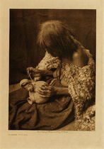 Edward S. Curtis -   Mohave Potter - Vintage Photogravure - Volume, 12.5 x 9.5 inches - The primitive Mohave would wear dresses of only willow bark cloth likely pictured here by Edward S. Curtis. Unlike the braids seen in many tribes the Mohave women would always wear their hair loose. For art the tribe was quite skilled at beadwork and some crude but lovely pottery. Pictured here is an interesting vessel with a small sculptural face. The Mohave woman is hard at work and does not look up at photographer Edward Curtis.
<br>
<br> This photogravure was taken in 1907 by Edward S. Curtis. The piece was printed on Deluxe Japanese Tissue and is available for sale in out Aspen Art Gallery.