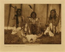 Edward S. Curtis -   Huka-Lowapi, The Altar Complete - Sioux - Vintage Photogravure - Volume, 9.5 x 12.5 inches - "At his bidding the Fire-carrier brings from the Singer's tipi and lays beside Work-do a bundle containing the four cherry sticks, the ear of corn, the eagle-feathers, the two Hunka pipes, and the red and blue paint — all to be used in preparing the altar. The bundle is passed through the sacred smoke. Returning to the Singer's tipi the Fire-carrier receives another skin, which he fills with sage and brings to the wakán-tipi. Spreading it between himself and the altar-space, Work-do calls for the buffalo-skull, which the Fire-carrier brings, wrapped in a buffalo-skin given him by the Singer, and lays tenderly on the sage. Work-do as cautiously removes the covering. He now thoroughly works a bit of red paint into a piece of buffalo-tallow, and purifies, first this mixture, then a roll of buffalo hair and a braid of sweet-grass. The hair and the sweet-grass he extends to the buffalo-skull, and after offering them to the spirits of the four quarters feigns to mark a line across its face from left temple to right, just above the orbits. After brushing the hair and grass downward over the face of the skull as if washing it, he offers the reddened tallow to the four spirits and paints a line across the forehead of the skull, with others down the right cheek, nose, and left cheek, and a few stripes on the horns. Back of the skull he plants the two forked sticks, lays the third wand across them, and leans the two Hunka pipes from the ground immediately in front of him to the cross-stick. Beneath it are laid two buffalo-skin rattles. The fourth stick with the ear of corn impaled on its tip and the eagle-feathers, one for each initiate, attached lower down, is implanted to the left of the two forked sticks and in a line with them. The altar is now complete." -Edward S. Curtis in "The North American Indian"
<br>
<br>This Edward Curtis photo was taken in 1907 of a completed ceremonial altar. Pictured are 3 Natives with a large buffalo skull in front of them. The piece was printed by Edward S. Curtis on Dutch Van Gelder and is available for sale in our Aspen Art Gallery.