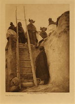Edward S. Curtis -   An Afternoon Chat - Vintage Photogravure - Volume, 12.5 x 9.5 inches - A handful of young Hopi maidens look down at the Edward Curtis’ camera from an adobe rooftop. The structure is of the typical Hopi adobe structure constructed of undressed stone fragments bound with mud plaster.  There is a large ladder leading up to the rooftop. The rooftops were made of wooden beams resting on the tops of the walls and covered in rod and grass thatching, a layer of plaster and then topped with a covering of dry earth. 
<br>
<br>These impressive structures can be up to four or even five stories high and are a fine show of ingenuity on the part of the Hopi people. The girls in this picture are dressed in costume, and most have their hair in two twisted buns on either side of their heads, indicating that they are unmarried. The ladder, which thins at the top bring the viewers eyes right up to the girls in the sky. This photogravure by Edward Curtis was taken in 1906 and is available at our Aspen Art Gallery.