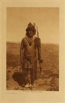 Edward S. Curtis - *50% OFF OPPORTUNITY* Numak-Mahana - Vintage Photogravure - Volume, 12.5 x 9.5 inches - In ancient times when Numak-mahana, the creator, was still dwelling among the Mandan, the people had a wonderful boat, which moved itself. It was called Idehe, "He Goes." The people would embark in it, and say, "Idehe, go!" and the boat would glide away across a certain body of water to the home of another people, where the Mandan obtained a kind of large shell called "the shell that has a noise." These strangers would feast until many of the visitors died. Numak-mahana, dissapproving of this, accompanied them on one of their voyages, disguising themselves, and running the long hollow stem of a water-lily through his body. Thus he was able to swallow all the food that was brought, and still call for more when the supply had been exhausted, for it passed through without lodging in his stomach. His hosts recognized him as a mahopini, and for the time abandoned the effort to kill their visitors. With seeming friendliness they promised to come to the Mandan the next year. Having returned home in safety, Numak-mahana at once planted a cedar post in the ground and painted it red. This, he said, would represent him to the people after his departure. Cedar was chosen because it is chief of all woods, outlasting all others. Around the post in a circle he erected wooden slabs, held together by two hoops of cottonwood saplings, one inside and one outside. "This," said he, "is minimi tahhe, and it shall be a breastwork for your protection. When the people from across the water come against you, they may kill some, but never all so long as this stands." In the following year their enemies came against them as expected, and caused great flood, but the hoops around minimi tahhe prevented the water from rising above them.