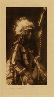Edward S. Curtis -   Good Lance - Ogalala - Vintage Photogravure - Volume, 12.5 x 9.5 inches - Born 1846. At thirteen he accompanied a party under his brother, then the possessor of the name Good Lance, against Pawnee, four woman and one man of whom they killed without loss to themselves. At twenty-five he led a party, and himself won first coup from the leader of the Pawnee, whom he struck with his lance and unhorsed, capturing the horse. On another occasion he struck with his coup stick a Pawnee boy left behind by his retreating companions; the father, armed with a Spencer rifle, turned to the rescue and fired at close range, but missed. Good Lance struck him with a sword, and others closing in killed father and son. He participated in ninety raids, mostly against the Pawnee. Fought against the troops in 1878, but was on the southern plains in 1876 and consequently took no part in the Custer fight. From a famous Cheyenne medicine-man south of the Platte he bought medicine of pulverized roots tied in deerskin, as a charm against bullets and arrows. The medicine-man clothed him in a buffalo-robe with horns and tail attached, and rubbed the charm all over his body. To prove that nothing could now harm him, the Cheyenne discharged a pistol at him, but the bullet only inflicted a skin wound in the arm; he then struck Good Lance in the back with a bayonet, but the resulting wound was slight. Good Lance paid the medicine-man a horse, and thereafter before entering a fight rubbed the medicine over his body. He fasted five times, the first time during two days and two nights. At dawn of the second day a white-breasted crow perched near him; at daylight the crow was transformed into a hawk. A thunder-storm approached and the hawk became a horse, which trotted toward the storm, while from beyond the ridge over which the animal passed was heard the sound of many horses whinnying. The storm broke, and holding his pipe close, he drew his robe over his crouching body and waited amid the rain and hail. After the storm abated he perceived that all the ground about him was perfectly dry, and that there were four men, each with a lance thrust into his body, who became mere ridges of hail. The faster concluded that the import of the vision was that he was destined to kill four men with a lance as he afterwards really did. His second fast was not rewarded, for he remained out only one day: the camp was being moved and he accompanied the others. On another occasion, he began to fast in the evening, standing upon a hilltop with his robe drawn about him. The following day as he slept under his robe he became aware of something creeping over his body toward his face. When he saw that it was a rattlesnake he was frightened; he dared not move. He set his teeth, and that thought came to him that this thing ought not to happen to one who had made offerings. The snake began to crawl away, and in his relief he moved; the reptile coiled at once and struck. Returning to the camp he went into a sweat-lodge, and though his body became stiff and swollen, he recovered.
<br>
<br>Provenance: 
<br>Art Institute of Chicago, Ryerson & Burnham Library