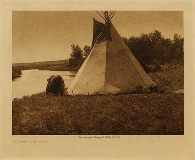 Edward S. Curtis - An Assiniboin Lodge - Vintage Photogravure - Volume, 9.5 x 12.5 inches - In this image an Assiniboin tipi rests on the shore of a river. A Native woman is bending over to stake the tipi and finish its construction. Tipis were easily portable so the people could move around for the change of seasons or to follow the buffalo. There is another tipi in the background that is only the under layer of wooden poles without the buffalo skin outer layer. This photogravure was taken by Edward S. Curtis in 1908 and is on display in our Aspen Art Gallery.
<br>
<br>Provenance: 
<br>Art Institute of Chicago, Ryerson & Burnham Library