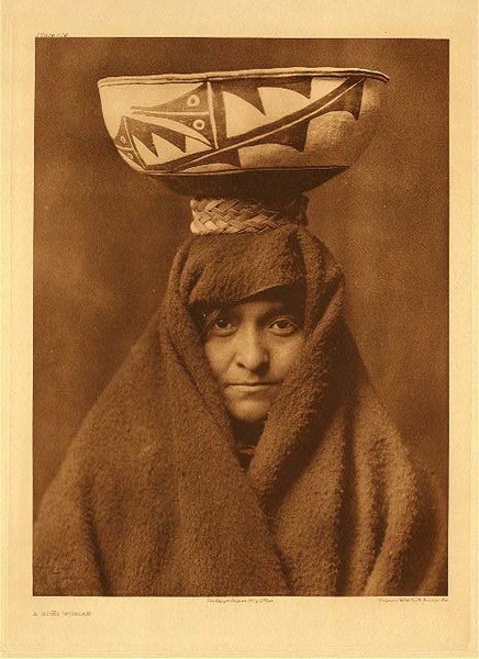 Edward S. Curtis - Plate 614 A Zuni Woman - Vintage Photogravure - Portfolio, 22 x 18 inches - This Zuni Woman seems fairly young and has chosen to have her face almost hidden in the draped brown material around her face and body. She looks directly into the camera and holds a large vessel upon her head. This vessel likely contains food for an intermission during a ceremony. A woven yucca ring helps her balance the large full vessel on her head without having it tip over. This photogravure was taken by Edward Curtis in 1903 and is available at our Aspen Art Gallery.