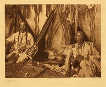 Edward S. Curtis -   Plate 188 In a Piegan Lodge - Vintage Photogravure - Portfolio, 18 x 22 inches - Description by Edward S. Curtis: <br> <br>Little Plume with his son Yellow Kidney occupies the position of honor, the space at the rear opposite the entrance. The picture is full of suggestion of the various Indian activities. In a prominent place lie the ever-present pipe and its accessories on the tobacco cutting-board. From the lodge-poles hang the buffalo-skin shield, the long medicine-bundle, an eagle-wing fan, and deerskin articles for accoutering the horse. The upper end of the rope is attached to the intersection of the lodge-poles, and in stormy weather the lower end is made fast to a stake near the centre of the floor space.<br><br>Desiring to keep the photographs as traditional and accurate as possible Curtis is accused of altering this photograph. It is said that he manipulated a clock out of the center of the piece. This is all the more interesting a photograph because of that, and showed Curtis' dedication to preserving this incredible group of people.