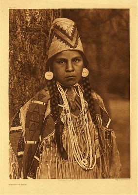 Edward S. Curtis -   Plate 269 Umatilla Maid - Vintage Photogravure - Portfolio, 22 x 18 inches - “Two distinct cultural areas are represented in the costume of this damsel. The familiar beadworked, deerskin dress is an acquisition from the plains culture, while the basketry hat and the shell-bead necklace hail from the pacific slope. Note the skin of the deer's tail fastened in front at the collar, as an aid in removing the garment.” – Edward Curtis.<br><br>It is curious to note that very little is written by Curtis regarding the Umatilla. The narrative in Volume VIII of The North American Indian, barely a paragraph speaks about them, only referring to a linguistic association to the Yakima.