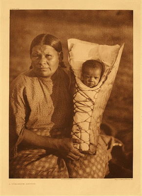Edward S. Curtis -   Plate 685 A Comanche Mother - Vintage Photogravure - Portfolio, 22 x 18 inches - In this image, Curtis candidly captures a Comanche Mother stoically holding a child in a cradle. The pair are pictured outside and in typical garb.<br><br>"The Comanche arer the sole representatives of the Shoshonean stock in Oklahoma. Like most of the tribes in that state, they were not natives of it, but were placed there for riddance. However, they differed from most other tribes removed to Oklahoma in that they were not strangers to the region, for it had been part of their hunting and raiding grounds for many years. No North American tribe ranged over so broad a territory. The Comanche were without prejudice in their selection of victims" from Edward S. Curtis' "The North American Indian", Volume XIX