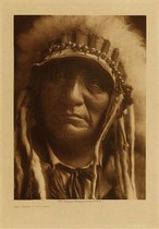 Edward S. Curtis -   Red Hawk - Ogalala (Same Individual as seen in "An Oasis in the Badlands") - Vintage Photogravure - Volume, 12.5 x 9.5 inches - Born 1854. First war-party in 1865 under Crazy Horse, against troops. Led an unsuccessful war-party at twenty-two against Shoshoni. First coup when twelve horse-raiding Blackfeet were discovered in a creek bottom and annihilated. Led another party against Shoshoni, but failed to find them; encountered and surrounded a white-horse troop. From a hill overlooking the fight Red Hawk saw soldiers dismount and charge. The Lakota fled, leaving him alone. A soldier came close and fired, but missed. Red Hawk did likewise, but while the soldier was reloading his carbine he fired again with his Winchester and heard a thump and "O-h-h-h!" A Cheyenne boy on horseback rushed in and struck the soldier, counting coup. Engaged in twenty battles, many with troops, among them the Custer fight of 1876; others with Pawnee, Apsaroke, Shoshoni, Cheyenne, and even with Sioux scouts.<br>Red Hawk fasted twice. The second time, after two days and a night, he saw a vision. As he slept, something from the west came galloping and panting. It circled about him, then went away. A voice said, "Look! I told you there would be many horses!" He looked, and saw a man holding green grass in his hand. Again the voice said, "There will be many horses about this season;" then he saw the speaker was a rose-hip, half red, half green. Then the creature went away and became a yellow-headed blackbird. It alighted on one of the offering poles, which bent as if under great weight. The bird became a man again, and said, "Look at this!" Red Hawk saw a village, into which the man threw two long-haired human heads. Said the voice,"I came to tell you something, and I have now told you. You have done right." Then the creature, becoming a bird, rose and disappeared in the south. Red Hawk slept, and heard a voice saying, "look at your village!" He saw four woman going around the village with their hair on the top of their heads, and their legs aflame. Following them was a naked man, mourning and singing the death-song.  Again he slept, and felt a hand on his head, shaking him, and as he awoke a voice said, "Arise, behold the face of your grandfather!" He looked to the eastward and saw the sun peeping above a ridge. The voice continued, "Listen! He is coming, anxious to eat." So he took his pipe and held the stem toward the rising sun. This time he knew he was not asleep, or dreaming: He knew he was on a hill three miles from the village. A few days later came news that of five who had gone against the enemy, four had been killed; one returned alive, and followed the four mourning wives around the camp singing the death-song. Still later they killed a Cheyenne and an Apsaroke scout, and the two heads were brought into camp.