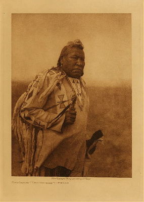 Edward S. Curtis -   Piksohkomi ("Calling - Bird") - Piegan - Vintage Photogravure - Volume, 12.5 x 9.5 inches - Piksohkomi (“Calling Bird”) appears in Edward S. Curtis’ eighteenth volume. From the Piegan tribe, this man is depicted wearing a beautiful deerskin shirt adorned with patterns (likely made by porcupine quills and beads) and weasel tails. A garment of this nature would be worn by most of the Plains Indian tribes. He is holding what appear to be religious items, with feathers. The arts and crafts of the Piegan Indians generally revolved around the production of clothing, shelters, and implements of war.<br><br>Edward S. Curtis took this photograph in 1926.