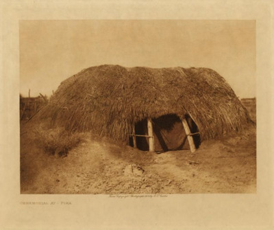 Edward S. Curtis -  *40% OFF OPPORTUNITY* Ceremonial Ki - Pima - Vintage Photogravure - Volume, 9.5 x 12.5 inches - The early dwellings of the Pima and their immediate congeners were quite alike: a dome-shape structure about seven feet high and fifteen feet in diameter at the base. A circular excavation twelve to eighteen inches deep was first made, in the centre of which four crotch posts were set about five feet apart, forming a square. Two heavy roof timbers rested upon these posts, five feet from the ground, supporting ten or more stout cross-beams. Numerous stave-like ribs of mesquite, tied to horizontal poles that extended around the outside like hoops, were stretched from the roof timbers to the bottom of the shallow excavation. The whole was thatched with arrow-brush and covered with clay, leaving only a small opening at the base on the eastern side as an entrance. Only a few of the old form of houses are now in use among the Pima. For the greater part their domiciles are rectangular, with flat roofs and straight walls of mud filled in between poles fastened horizontally to opposite sides of stout posts, or with brush-wattled walls plastered inside and out with mud.